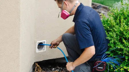 Zerorez Technician wearing a respirator mask kneeling outdoors feeding blue cables into a wall opening near a shrub.