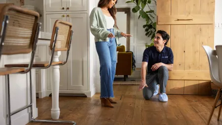 Woman in jeans and man in navy shirt discuss wood floor quality in modern kitchen with natural light.