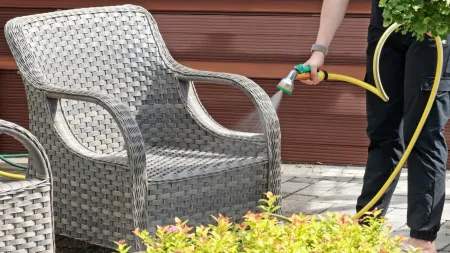 A person cleaning a wicker chair with a garden hose in a sunny outdoor setting.