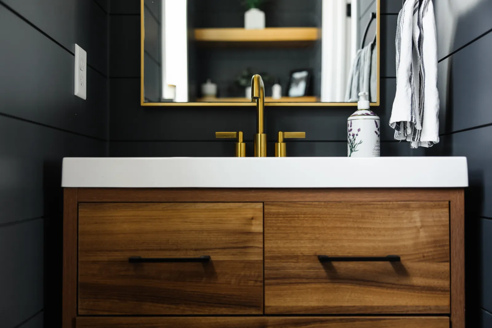 Wood cabinets/drawer below white countertop sink in a black shiplap bathroom with a hand towel hanging