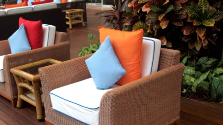 Outdoor wicker chairs with blue and orange pillows on wooden deck next to green plants and small table